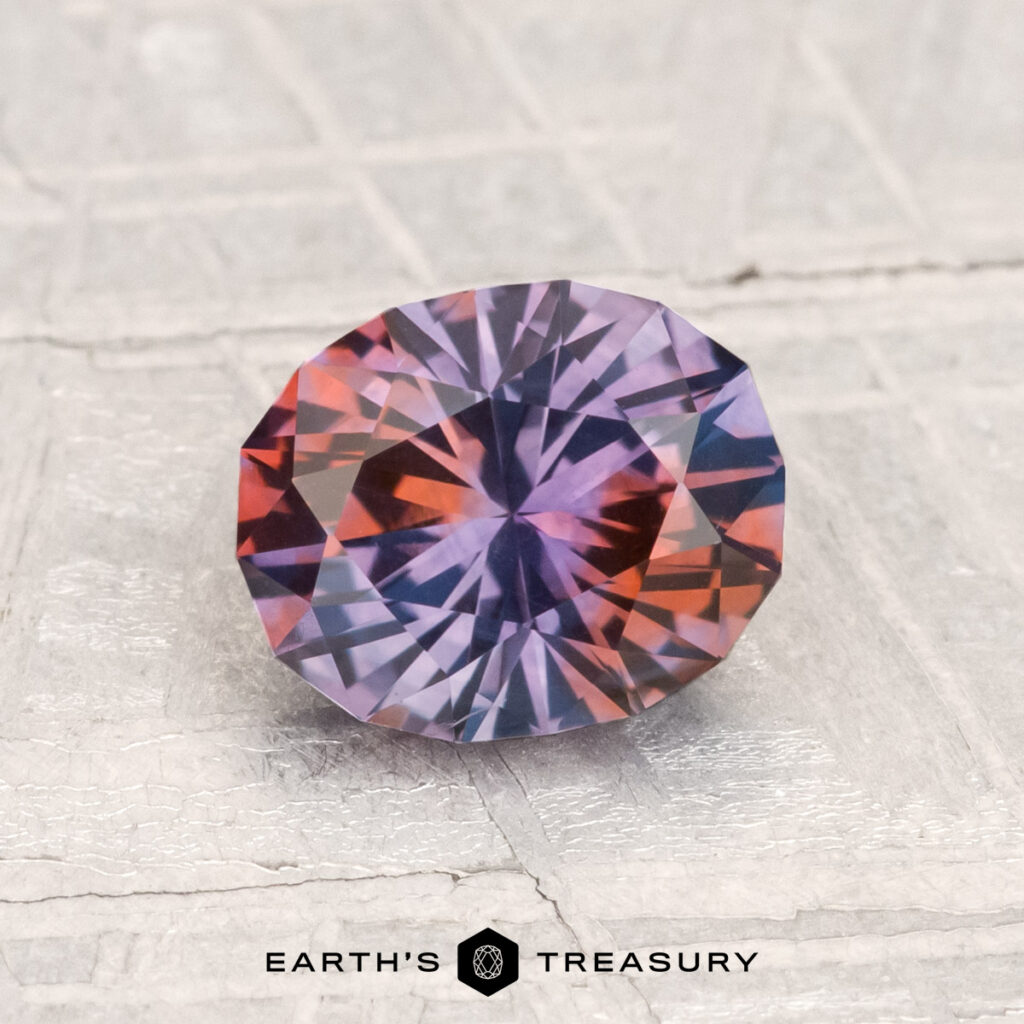 A faceted, oval-cut 1.58-Carat Montana Sapphire (Heated) with a blend of purple and orange hues rests on a textured light surface. "Earth's Treasury" appears at the bottom.
