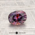 A faceted, oval 1.42-carat heated Montana sapphire with red and purple hues rests on a textured silver-gray surface; the "EARTH'S TREASURY" logo appears at the bottom.
