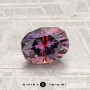 A faceted, oval 1.42-carat heated Montana sapphire with red and purple hues rests on a textured silver-gray surface; the "EARTH'S TREASURY" logo appears at the bottom.