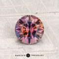A 1.45-carat heated Montana sapphire with a pinkish-purple, round faceted shape is shown on a textured surface, featuring "EARTH'S TREASURY" at the bottom.
