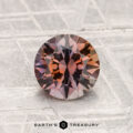 A 1.45-carat heated Montana sapphire with reddish-brown and purple tones is displayed on a textured metallic background featuring the text “EARTH’S TREASURY” at the bottom.