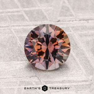 A 1.45-carat heated Montana sapphire with reddish-brown and purple tones is displayed on a textured metallic background featuring the text “EARTH’S TREASURY” at the bottom.