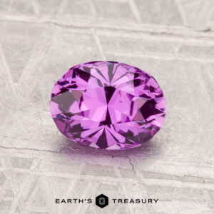 A 1.29-carat, oval-cut Madagascar sapphire is displayed on a textured light gray surface with the logo and text "EARTH'S TREASURY" below.
