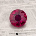 A round, faceted 1.26-carat heated ruby is displayed on a textured gray surface with the text "EARTH'S TREASURY" at the bottom.