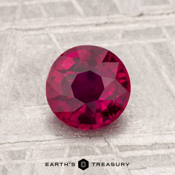 A round, faceted 1.26-carat heated ruby is displayed on a textured gray surface with the text "EARTH'S TREASURY" at the bottom.