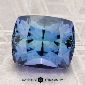 A 6.30-carat unheated tanzanite (zoisite) with stunning internal reflections rests on a textured grey surface, with "EARTH'S TREASURY" displayed below.