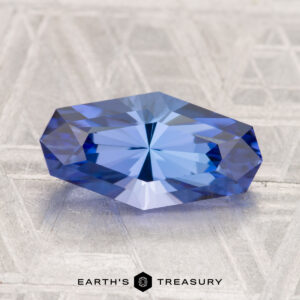 A 5.54-carat unheated tanzanite (zoisite) with geometric facets rests on a textured light gray surface; the "EARTH'S TREASURY" logo appears at the bottom.