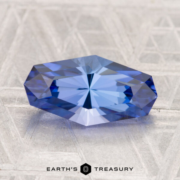 5.54-Carat Unheated Tanzanite (Zoisite) A 5.54-carat unheated tanzanite (zoisite) with geometric facets rests on a textured light gray surface; the "EARTH'S TREASURY" logo appears at the bottom.