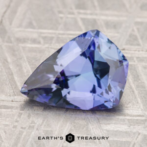 A 5.39-carat unheated tanzanite (zoisite) gemstone with a bluish-purple hue rests on a textured gray surface, with "EARTH'S TREASURY" at the bottom.