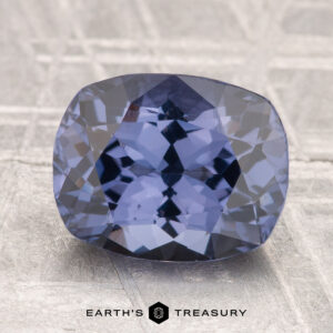 A faceted, cushion-cut 3.33-carat blue Spinel rests on a light, textured surface with the "Earth's Treasury" logo below.