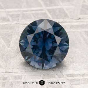 A 2.12-carat heated Montana sapphire, round and faceted with a blue hue, rests on a textured light surface. "EARTH'S TREASURY" is shown at the image's bottom.