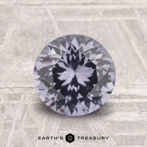 A 1.79-carat round brilliant-cut spinel with a lavender hue rests on a metallic surface with etched patterns. "EARTH'S TREASURY" is displayed at the bottom.