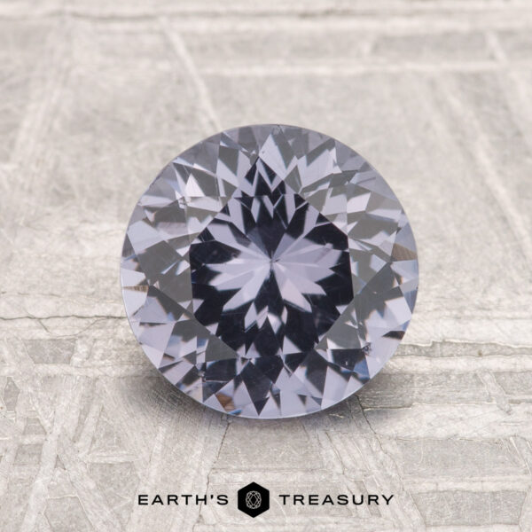 1.79-Carat Spinel A 1.79-carat round brilliant-cut spinel with a lavender hue rests on a metallic surface with etched patterns. "EARTH'S TREASURY" is displayed at the bottom.