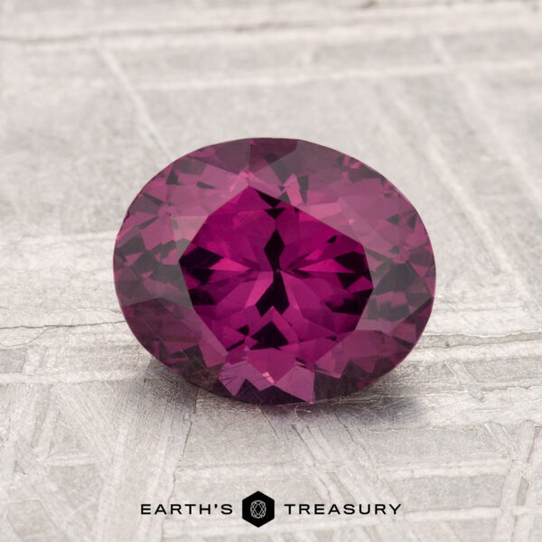 1.65-Carat Spinel A 1.65-carat faceted oval-cut pinkish-purple spinel is displayed on a light, textured surface with the text "EARTH'S TREASURY" below.