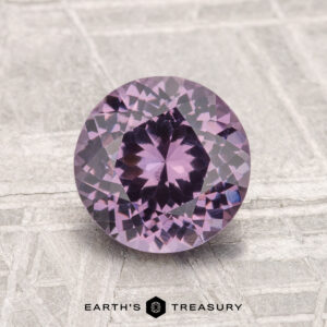 A 1.57-carat round, faceted purple spinel gemstone displayed on a textured gray surface with the text "EARTH'S TREASURY" at the bottom.