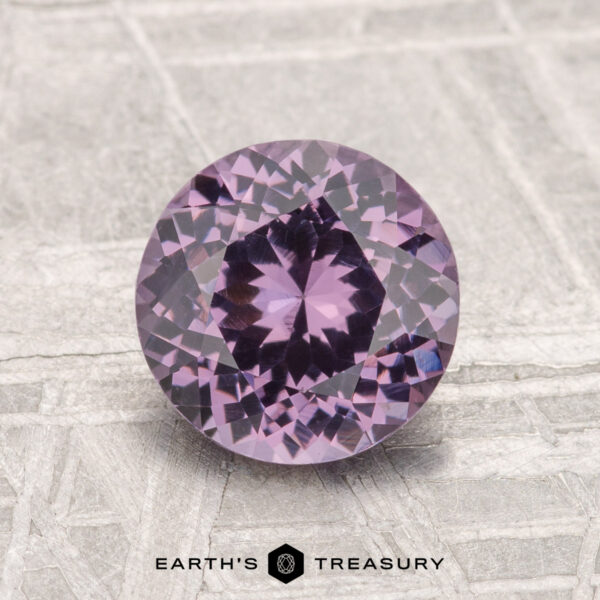 1.57-Carat Spinel A 1.57-carat round, faceted purple spinel gemstone displayed on a textured gray surface with the text "EARTH'S TREASURY" at the bottom.