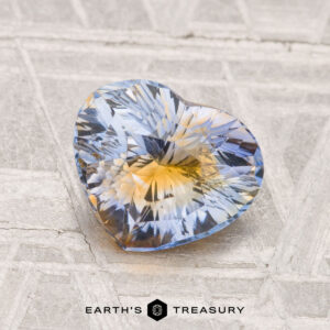 A 1.86-carat, heart-shaped sapphire with blue and yellow hues rests on a textured surface. "EARTH'S TREASURY" appears below the gemstone.