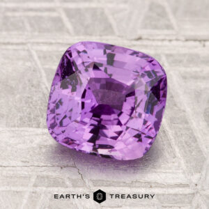 A 2.51-carat faceted purple sapphire rests on a textured metallic surface, with "EARTH'S TREASURY" displayed at the bottom.
