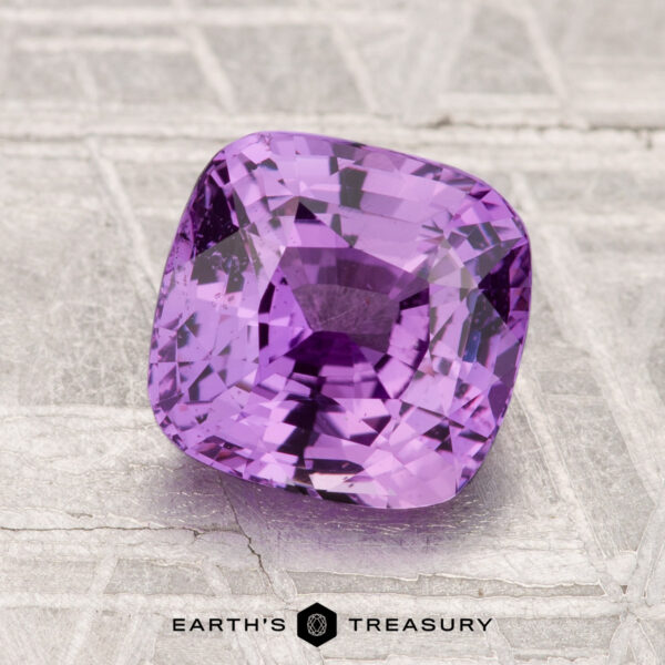 2.51-Carat Sapphire A 2.51-carat faceted purple sapphire rests on a textured metallic surface, with "EARTH'S TREASURY" displayed at the bottom.