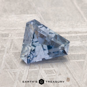 A 1.24-carat faceted blue sapphire in a triangular shape is displayed on a textured, light surface with the Earth's Treasury logo at the bottom.