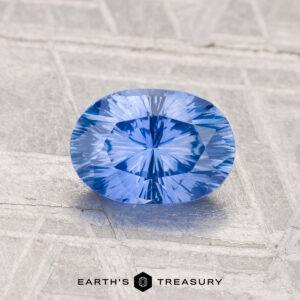 A 1.24-carat heated Ceylon sapphire, faceted in an oval shape, rests on a textured gray surface with the "Earth's Treasury" logo at the bottom.