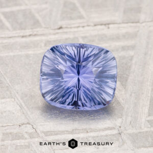 A 1.34-carat Ceylon sapphire with a cushion cut is shown on a textured gray surface, featuring the "Earth's Treasury" logo at the bottom.