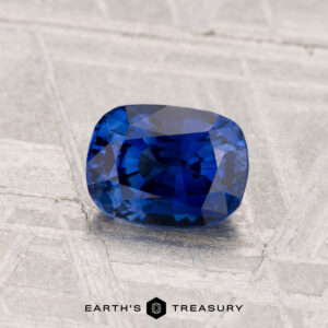 A 1.18-carat heated Ceylon sapphire, cushion-cut and deep blue, is displayed on a textured gray surface with the text "EARTH'S TREASURY" below.