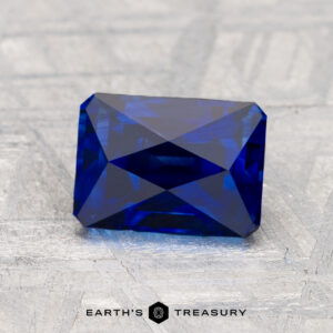 A rectangular, deep blue faceted 2.21-carat sapphire is displayed on a textured surface with the "Earth's Treasury" logo beneath it.