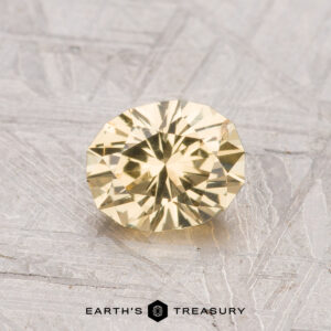 A 0.99-carat round, faceted grossular garnet with a yellow hue is displayed on a gray textured surface, with the text "EARTH'S TREASURY" at the bottom.