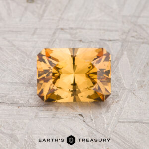 A 1.11-carat rectangular step-cut golden-yellow Grossular Garnet rests on a textured gray surface, with "EARTH'S TREASURY" text and logo at the bottom.