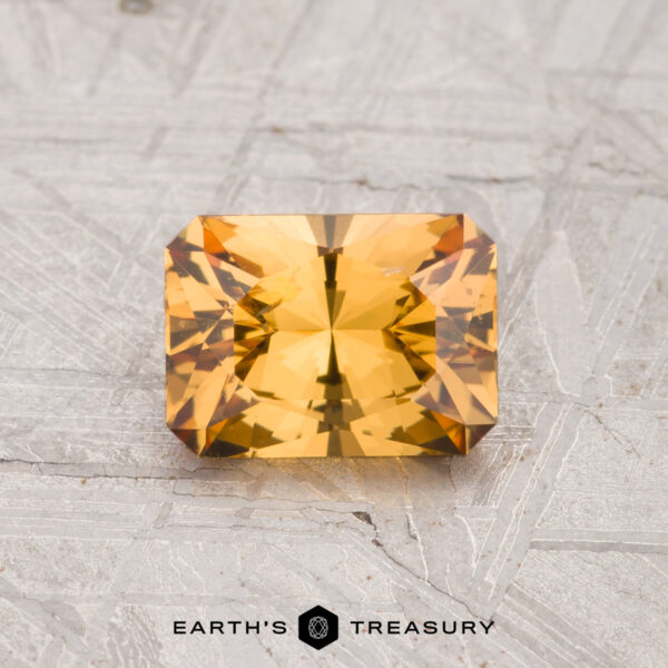 1.11-Carat Grossular Garnet A 1.11-carat rectangular step-cut golden-yellow Grossular Garnet rests on a textured gray surface, with "EARTH'S TREASURY" text and logo at the bottom.