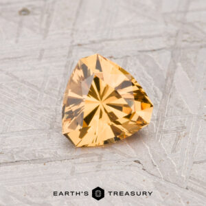 A 0.65-carat grossular garnet, cut in a triangular shape and yellow in color, rests on a textured gray surface with "Earth's Treasury" displayed below.