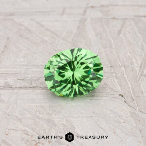 A 0.45-carat Tsavorite Garnet, cut in a round brilliant style, is shown on a textured gray surface with the "Earth's Treasury" logo beneath it.