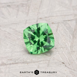 A 0.55-carat cushion-cut Tsavorite garnet rests on a textured gray surface, with “EARTH'S TREASURY” displayed at the bottom.
