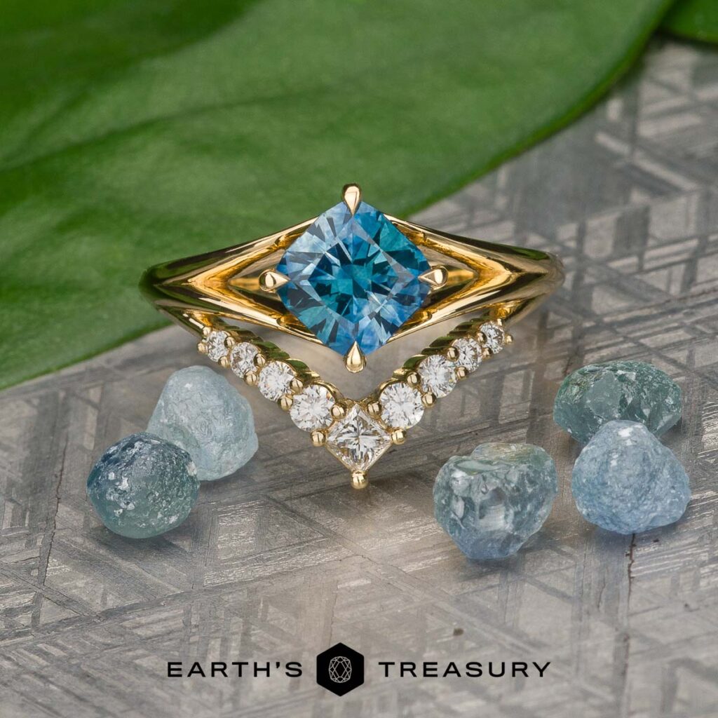 Product 1, a gold ring featuring a square blue gemstone and a V-shaped diamond band, is displayed among rough blue stones on a patterned surface.