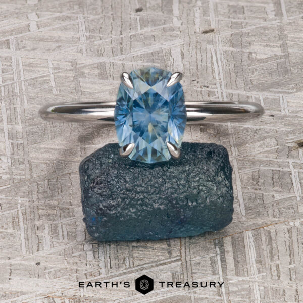 The "Tiara" ring in 18k white gold with 2.85-carat Montana sapphire The "Tiara" ring in 18k white gold, featuring a 2.85-carat Montana sapphire, is displayed on a rough blue stone background with the Earth's Treasury logo at the bottom.