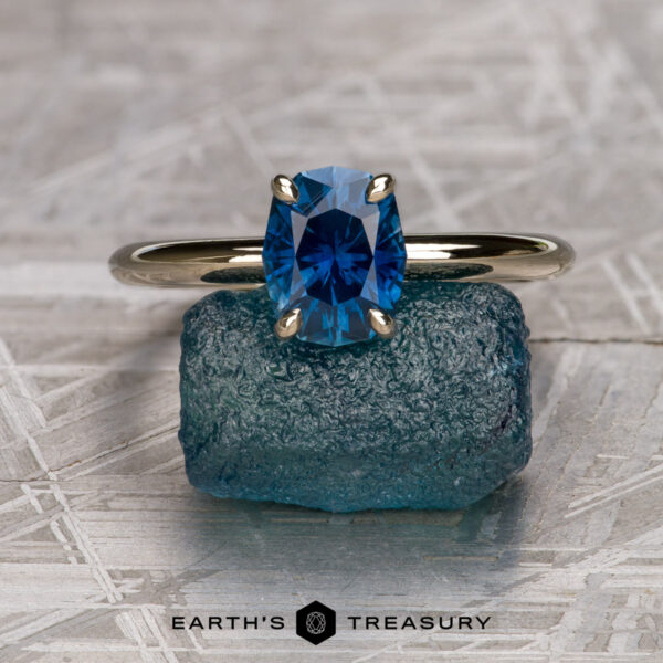 The "Tiara" ring in 14K unplated white gold with 1.37-carat Montana sapphire The "Tiara" ring in 14K unplated white gold, featuring a 1.37-carat Montana sapphire, is displayed upright on a rough blue crystal with the Earth's Treasury logo at the bottom.