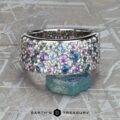 The Wide “Borealis” Ring in Blue-Violet Sapphire, featuring clustered blue, purple, and clear gemstones, is displayed upright on a rough blue stone with a textured gray background.