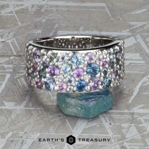 The Wide “Borealis” Ring in Blue-Violet Sapphire, featuring clustered blue, purple, and clear gemstones, is displayed upright on a rough blue stone with a textured gray background.