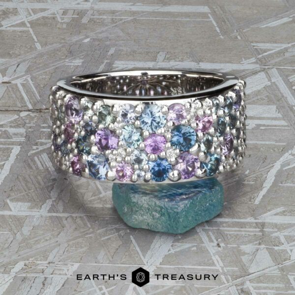 The Wide “Borealis” Ring in Blue-Violet Sapphire The Wide “Borealis” Ring in Blue-Violet Sapphire, featuring clustered blue, purple, and clear gemstones, is displayed upright on a rough blue stone with a textured gray background.