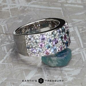 The Wide “Borealis” Ring in Blue-Violet Sapphire rests on a rough blue-green crystal, featuring vibrant gemstones and the Earth's Treasury logo below.
