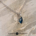 The "Brisa" pendant in 14K white gold features a 1.25-carat marquise-cut Montana sapphire on a silver chain, displayed on light wood. "EARTH'S TREASURY" logo appears at the bottom.