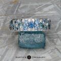The Thin "Borealis" ring in 14k white gold, set with blue and teal gemstones, rests atop a rough blue crystal on a textured gray surface. The Earth's Treasury logo appears at the bottom.