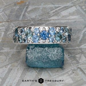 The Thin "Borealis" ring in 14k white gold, set with blue and teal gemstones, rests atop a rough blue crystal on a textured gray surface. The Earth's Treasury logo appears at the bottom.