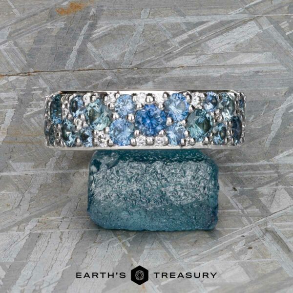 The Thin "Borealis" ring in 14k white gold The Thin "Borealis" ring in 14k white gold, set with blue and teal gemstones, rests atop a rough blue crystal on a textured gray surface. The Earth's Treasury logo appears at the bottom.
