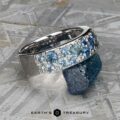 The Thin "Borealis" ring in 14k white gold, adorned with blue and white gemstones, is shown resting on a rough blue stone atop a textured metallic surface.