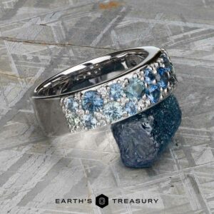The Thin "Borealis" ring in 14k white gold, adorned with blue and white gemstones, is shown resting on a rough blue stone atop a textured metallic surface.
