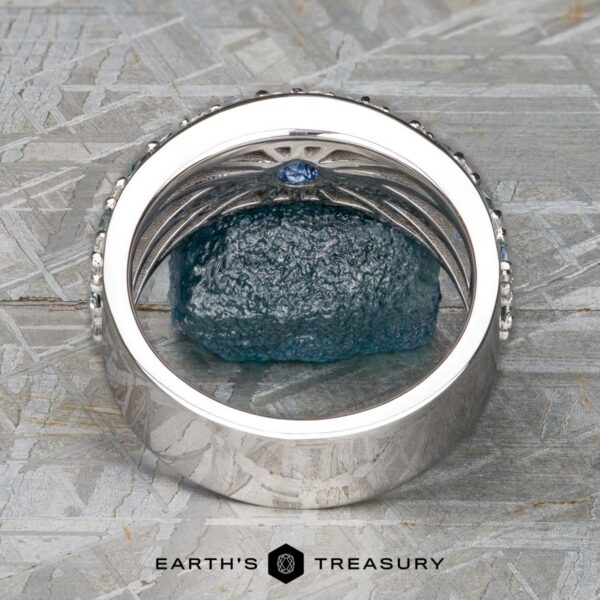 The Thin "Borealis" ring in 14k white gold The Thin "Borealis" ring in 14k white gold features an intricate design and a small blue gemstone, displayed upright on a rough blue mineral with a geometric backdrop and the “Earth’s Treasury” logo below.