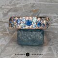 The Thin "Borealis" ring in 14k rose gold, featuring blue and teal gemstones, is displayed balanced on a rough blue-green mineral atop a textured metallic surface.