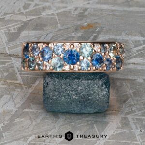 The Thin "Borealis" ring in 14k rose gold, featuring blue and teal gemstones, is displayed balanced on a rough blue-green mineral atop a textured metallic surface.